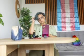 A transgender man sits at a wooden table, engaging in a makeup tutorial using his mobile phone.