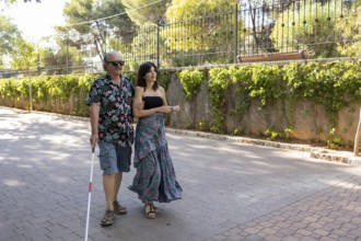 Photograph capturing a blind man using a white cane, being guided through town by his supportive