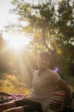 A young man relaxes outdoors by a lake, smiling broadly as he enjoys the warm summer sunshine
