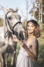 Young woman in a white dress gently pets a white horse on a sunny day. The serene outdoor setting