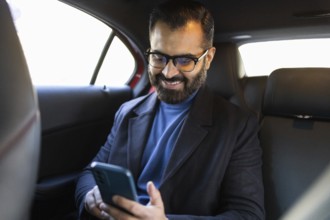 A happy Indian man, looking down at his smartphone, sits in a car, dressed in a winter coat and