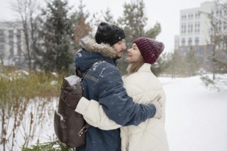 A couple warmly embraces in a snowy outdoor setting, surrounded by trees They wear winter attire