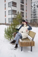 A couple sits on a wooden bench surrounded by snow, holding a small Christmas fir They are bundled