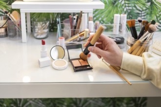 A hand uses a brush to apply organic makeup on a white vanity Various cosmetics, including lipstick