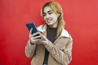 A Mixed-race woman smiles as she uses a smartphone, against a vibrant red background She is wearing