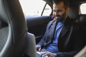 A happy Indian man dressed in professional attire works on his laptop while sitting in the backseat