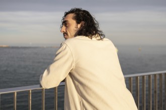 A man with long hair stands by a railing, gazing at the serene ocean under soft sunlight, conveying