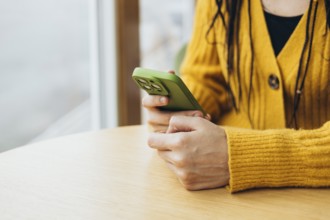 A person in a cozy yellow sweater holds a green smartphone while sitting at a wooden table near a
