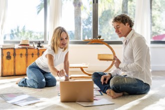 A man and woman sitting on the floor at home, engaging in work and planning activities. The man and