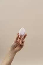 A hand gracefully holds a teardrop-shaped cosmetic sponge against a soft beige background,