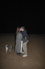 A couple shares a tender kiss on a dark beach night, their small dog beside them The scene captures