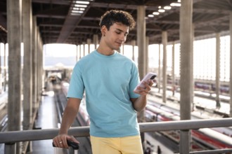 A young man in a blue t-shirt stands at a train station, holding a suitcase and using his