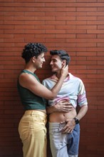 A joyful gay couple embracing and smiling at each other, standing against a red brick wall. The