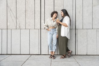A relaxed lesbian couple enjoys a casual moment together, leaning against a modern stone wall. Both