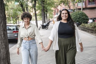 A lesbian couple is walking hand in hand on a city street, smiling and enjoying a sunny day. The