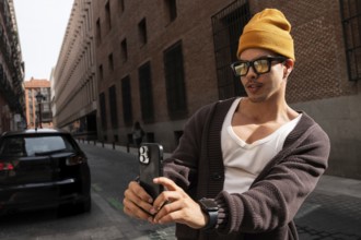 A stylish latin man wearing glasses and a yellow beanie takes a picture with his smartphone on a