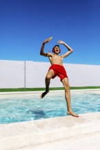 A joyful young teenage boy in red trunks leaps into a pool, wearing headphones and holding a