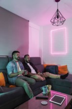A gay couple relaxes on a sofa, playing video games in a warmly lit living space with modern decor.