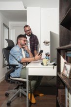 A gay couple collaborates at home, with one seated and the other standing, reviewing a smartphone.