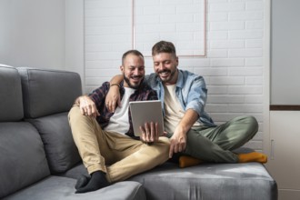 A joyful gay couple is comfortably seated on a sofa, engaged with a tablet. Their smiles reflect