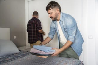 A gay couple organizes clothes and tidies up their bedroom. One is folding laundry on the bed while