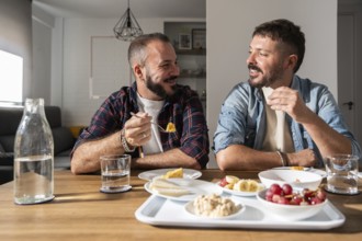 A happy gay couple sits together in a bright dining room, sharing a meal and enjoying each other's