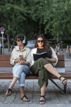 Lesbian couple relaxes on a park bench. One is reading a book, while the other listens to music,