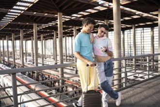 A joyful gay couple at a modern train station, sharing a moment with a smartphone, ready for their