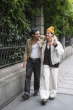 A joyful gay couple strolls arm in arm along a leafy city sidewalk, sharing smiles and enjoying
