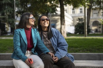 A latin lesbian couple sitting on a bench, smiling and holding hands. They wear sunglasses and