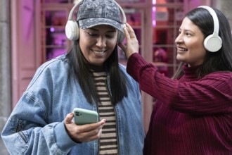 A smiling Latin lesbian couple enjoying music together with headphones. One holds a smartphone,