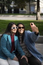 A smiling Latin lesbian couple enjoys a sunny day at the park, embracing while taking a selfie with