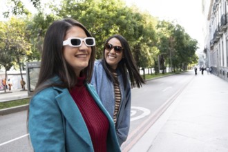 A joyful Latin lesbian couple strolls hand in hand down a tree-lined city street. They wear stylish