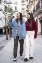 A joyful latin lesbian couple enjoys a sunny stroll, capturing memories on a city street. They