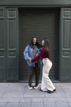A cheerful moment between latin lesbian couple standing on a sidewalk in front of a green shutter,