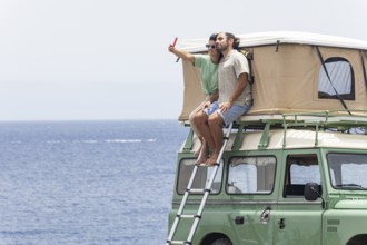 A blind man and a woman sit on the rooftop tent of a motorhome, enjoying a coastal view while