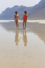 Lesbians walk hand in hand along a pristine beach, their reflections in the wet sand creating