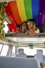 A joyful lesbian couple relaxes in a van under a rainbow flag, sharing laughs and browsing on a