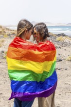 Two women wrapped in a rainbow flag share a moment on a rocky beach, symbolizing love and pride in