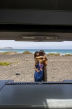 A lesbian couple shares an intimate embrace by the ocean, viewed from inside a camper. The serene