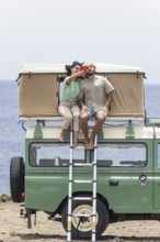 A blind man and a woman sit on the rooftop tent of a motorhome, enjoying a coastal view while