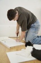 A man assembles new furniture on a hardwood floor with a curious cat watching nearby. He uses tools