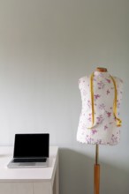 A floral-patterned tailor's mannequin with a yellow measuring tape stands next to a desk with a