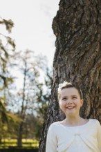 A joyful girl in a white First Communion dress stands beside a tree, beaming with happiness.