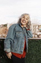 A joyful woman with white hair and a denim jacket smiles brightly while standing on her balcony,