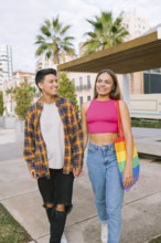 A happy lesbian couple walks hand in hand through an urban park. One partner carries a Pride flag