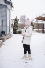 A girl dressed warmly in a pink hat and white coat enjoys winter games in the snow outside her home