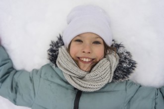 A smiling child lies in the snow, wearing a warm jacket, white hat, and knitted scarf. The joy and