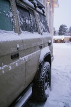 A snow-covered vehicle is parked near a warmly lit building in a snowy setting. The scene captures