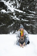 A woman in a winter coat kneels with a dog in a snow-covered forest. The serene winter landscape